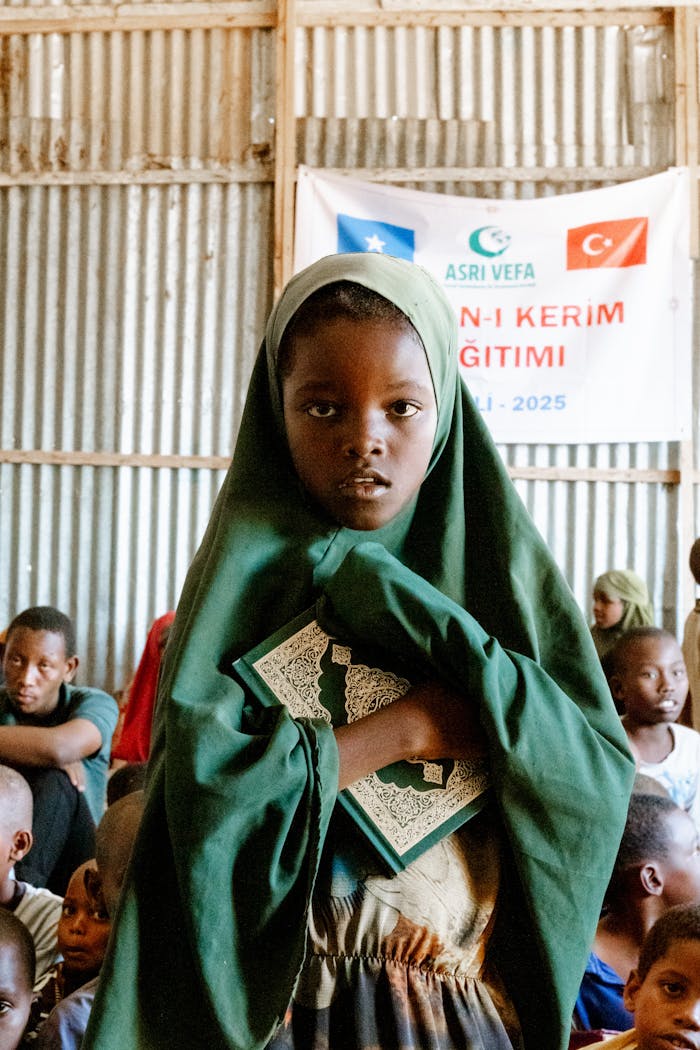 A young girl in a green hijab holds a religious book in an educational setting.